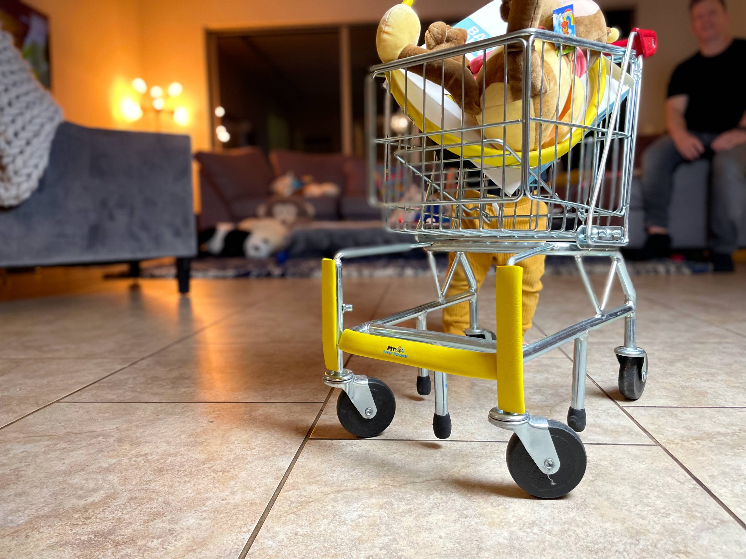6 months old baby pushing a toy shopping cart that is wrapped with the yellow baby bumper and is filled with a stuffed animal. We see the Mr B's Baby Bumper logo on the front of the bumper and shopping cart.